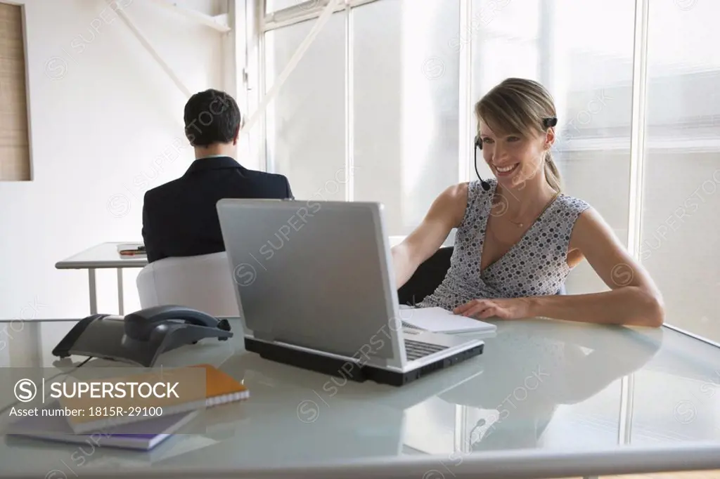 Business woman with headset working on laptop, male colleague in background