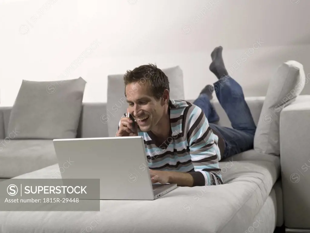 Man lying on sofa, using laptop and mobile phone