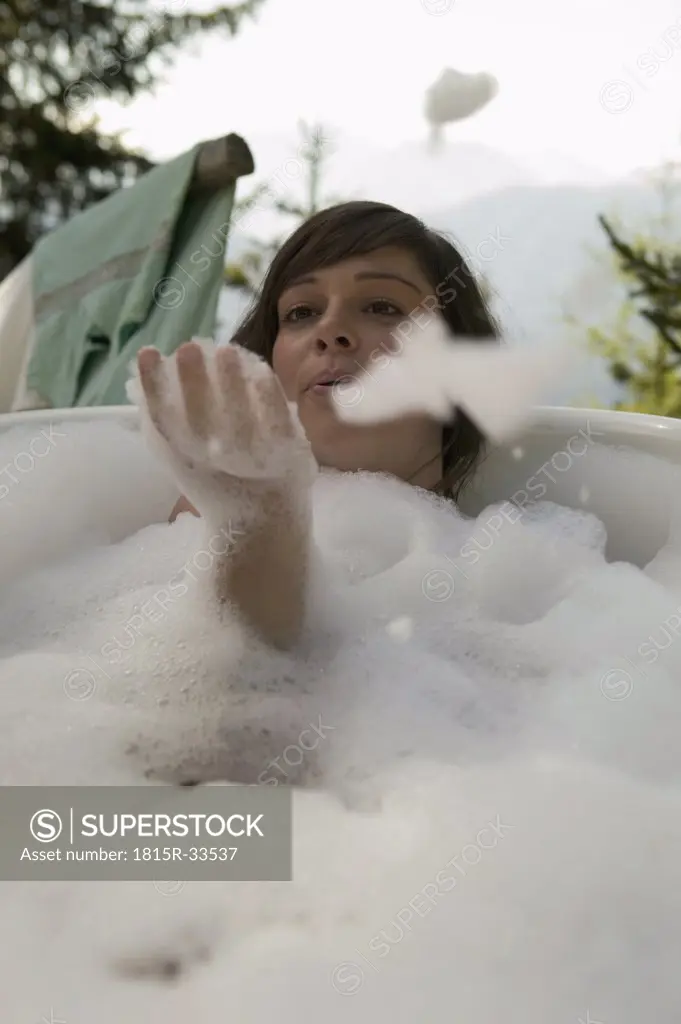Young woman lying in bathtub, blowing suds, outdoors
