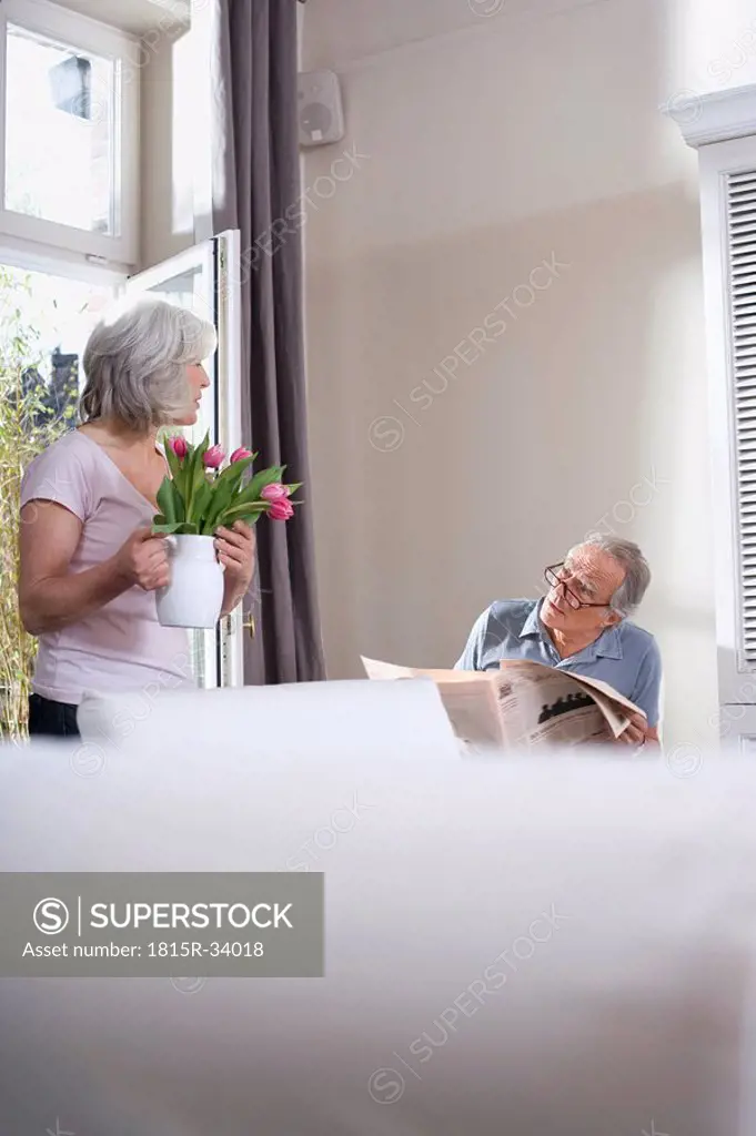 Senior woman holding flower vase, man reading newspaper