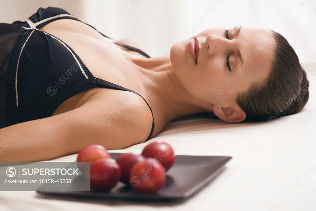 Young woman wearing neglige, relaxing on bed alongside tray with plums