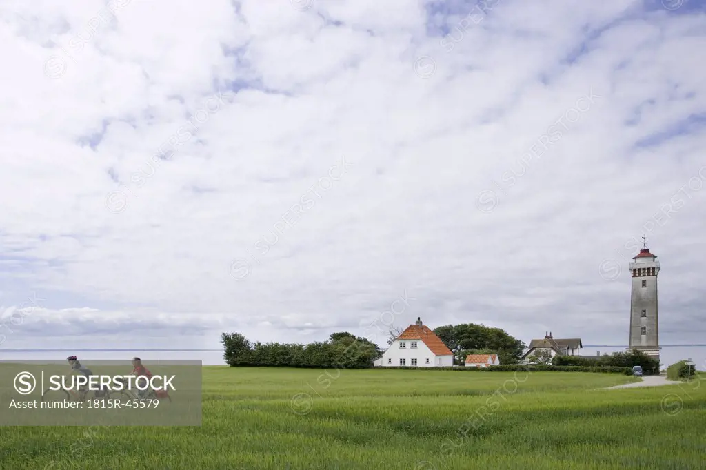 Denmark, Fuenen, Couple mountain biking across farm track, lighthouse in background