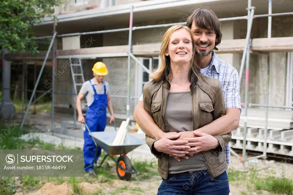 Young couple at site embracing, construction worker in background
