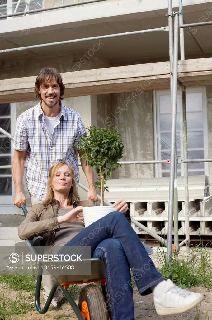 Young couple at construction site, woman sitting in wheelbarrow