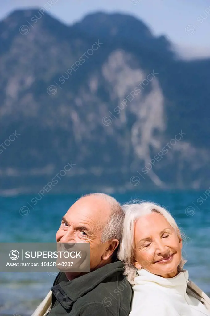 Germany, Bavaria, Senior couple relaxing on lakeshore, sitting back to back, smiling, portrait