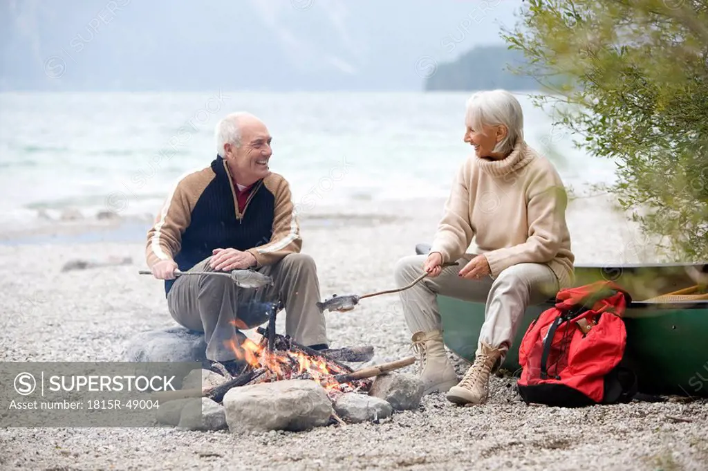 Germany, Bavaria, Walchensee, Senior couple sitting at campfire, grilling fish