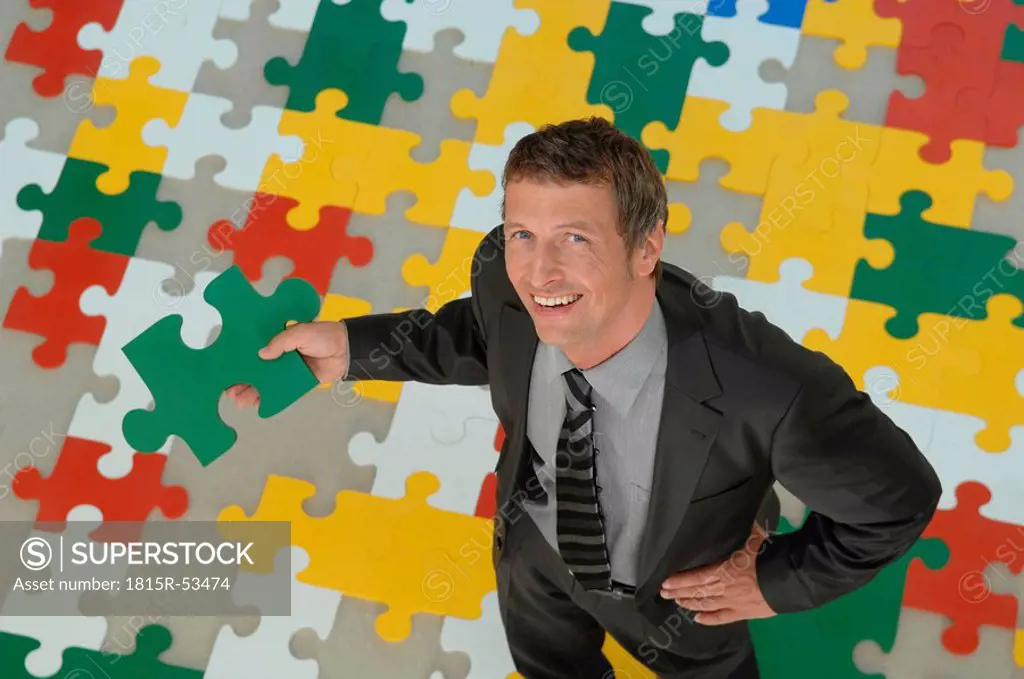 Businessman standing on jigsaw puzzle, holding piece of puzzle, smiling, portrait, elevated view