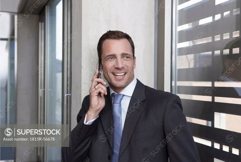 Germany, Cologne, Businessman leaning against wall using mobile phone, smiling, portrait