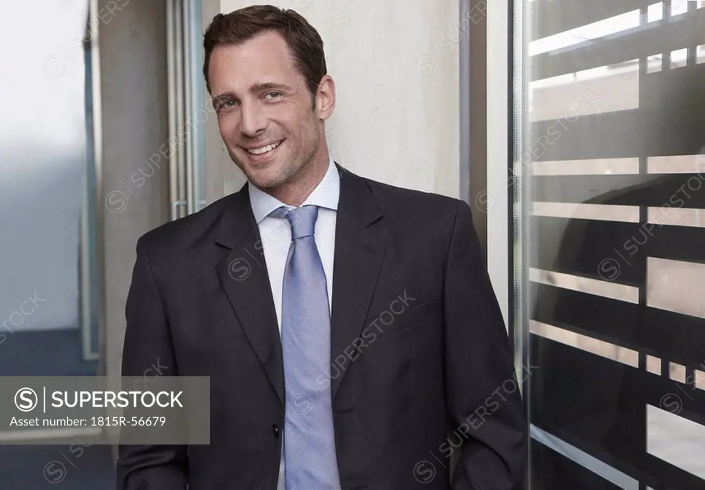Germany, Cologne, Businessman standing in corridor leaning against wall, smiling, portrait