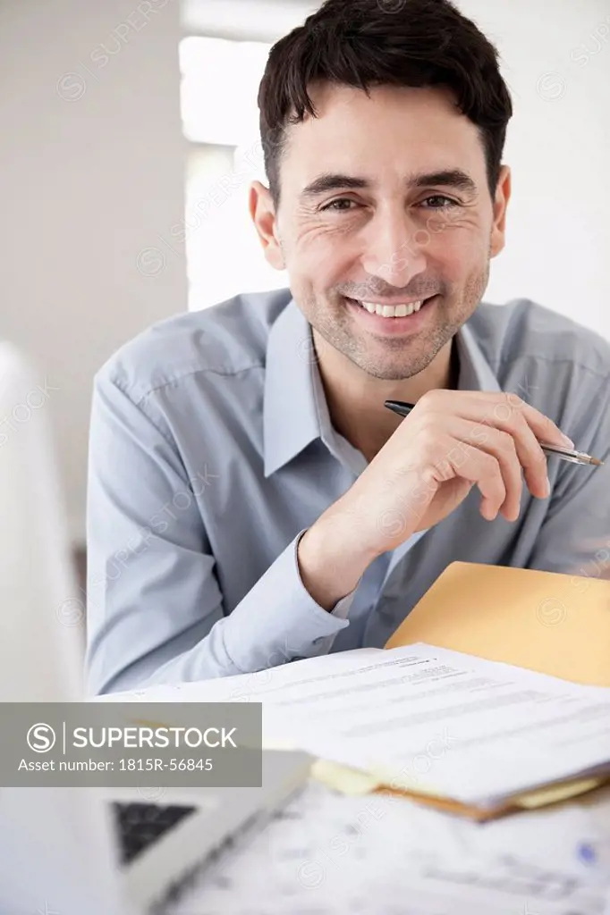 Businessman in office holding ballpen, smiling, portrait