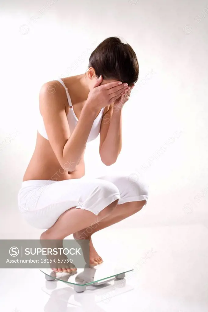 Young woman squatting on scales, hands to heas