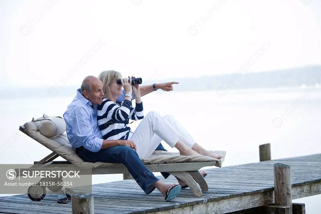 Germany, Bavaria, Starnberger See, Senior couple sitting on sunlounger, woman looking through binocular