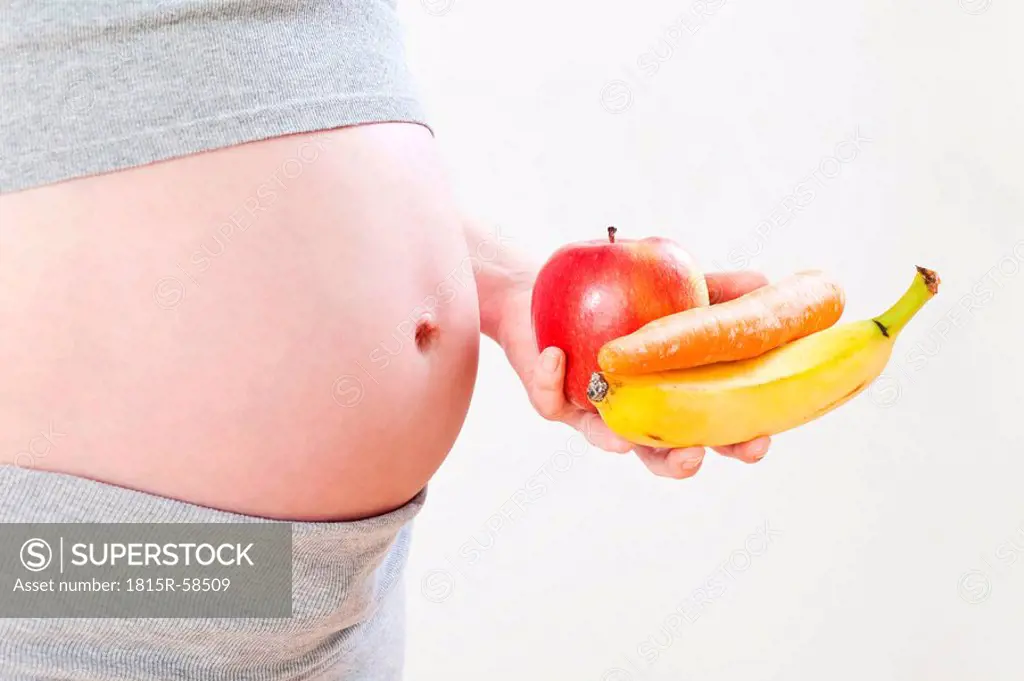 Pregnant woman holding banana, apple and carrot, mid section, close_up