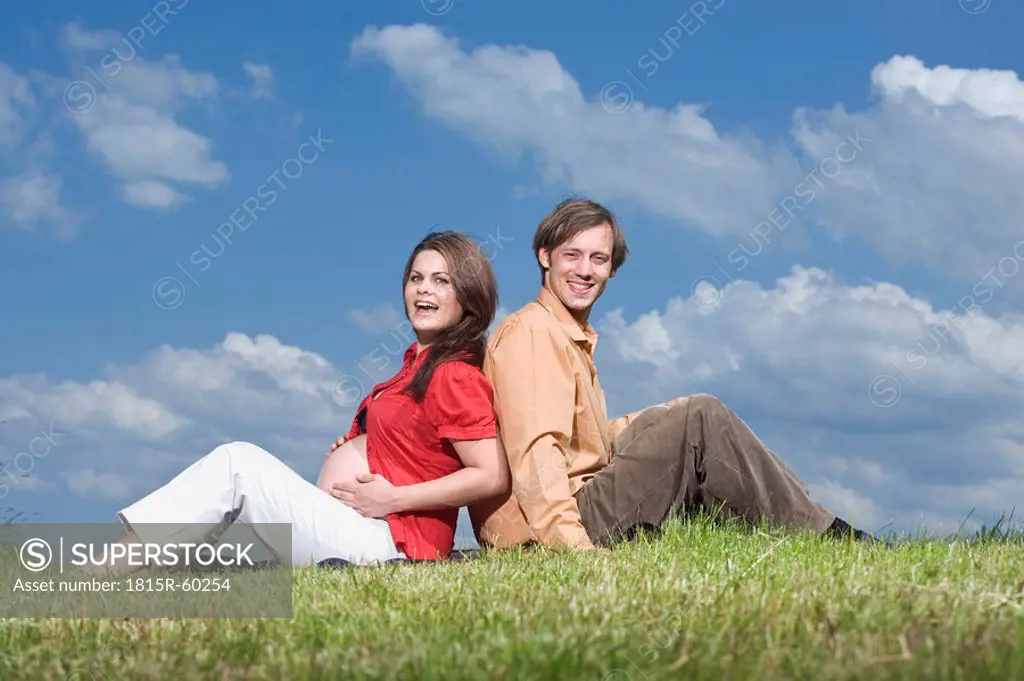 Couple sitting back to back in meadow, smiling, portrait