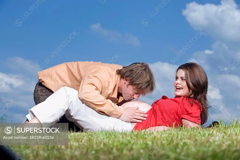 Woman lying in meadow, man kissing belly of pregnant woman, portrait