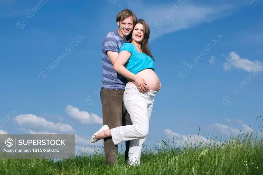 Young man embracing pregnant woman, standing in meadow, smiling, portrait