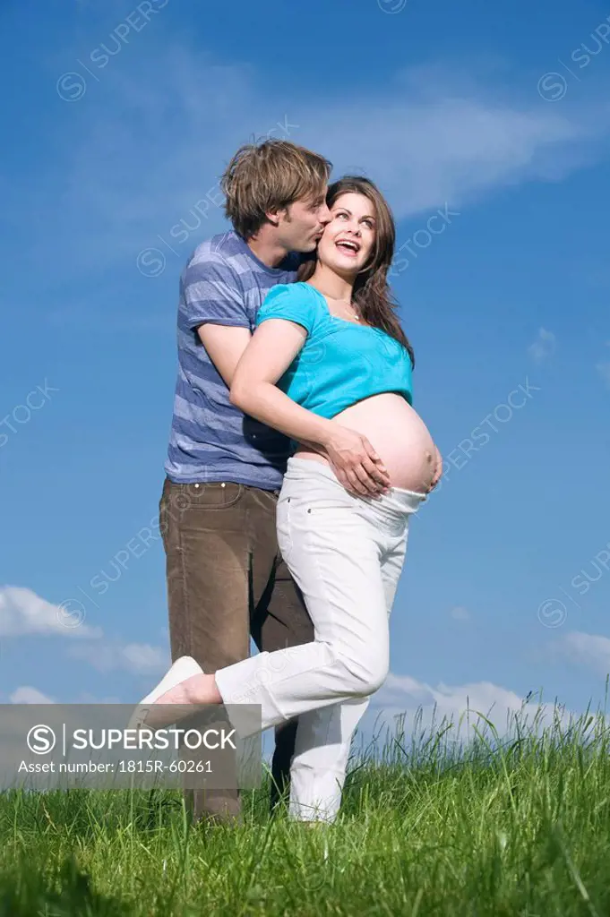Young man embracing pregnant woman, standing in meadow, smiling, portrait