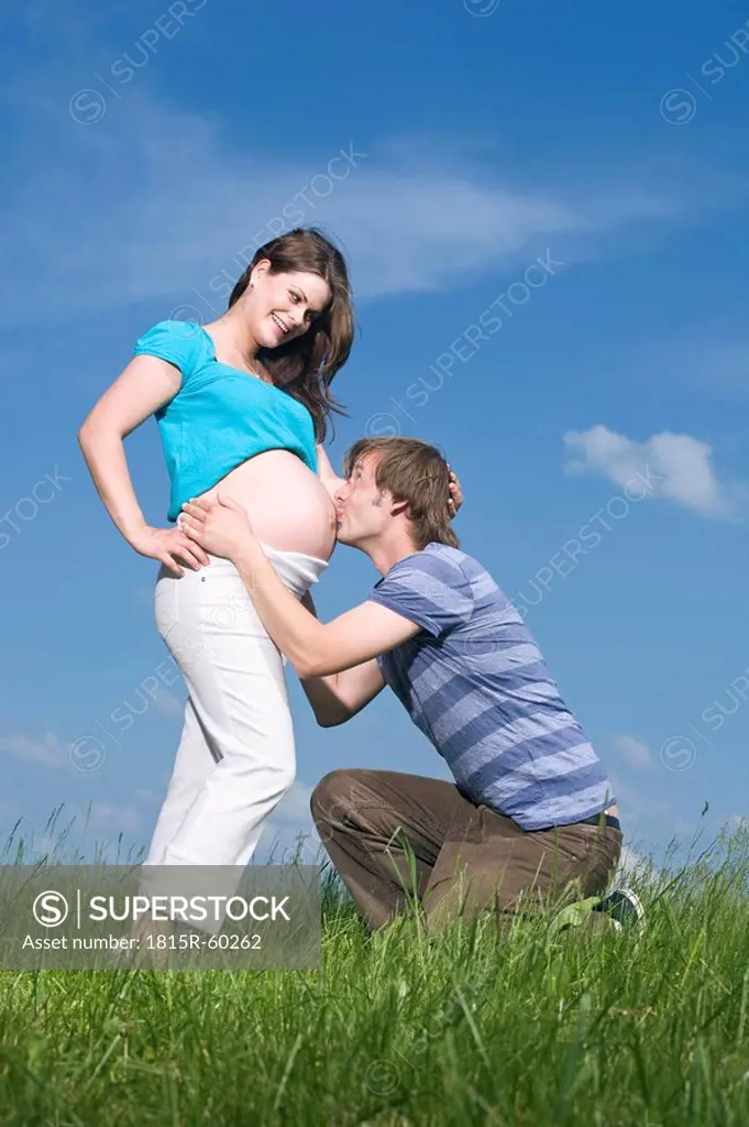 Woman standing in meadow, man kissing belly of pregnant woman, portrait