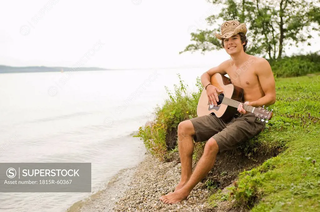 Germany, Bavaria, Starnberger See, Young man sits on lakeshore playing guitar, smiling, portrait