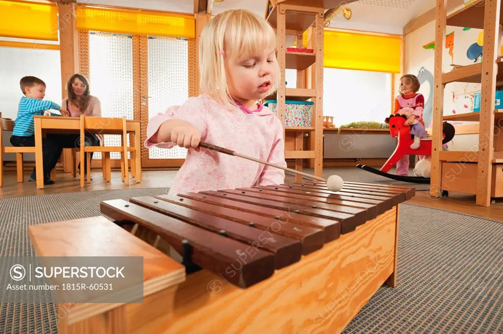 Germany, Girl 3_4 playing xylophone, Female nursery teacher with children in background, portrait