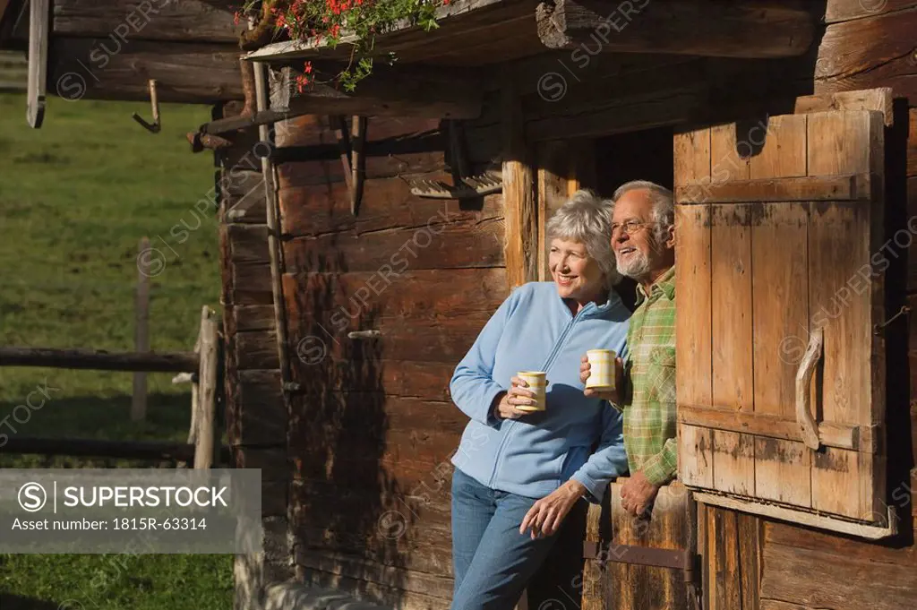 Austria, Karwedel, Senior couple leaning on log cabin, holding mugs