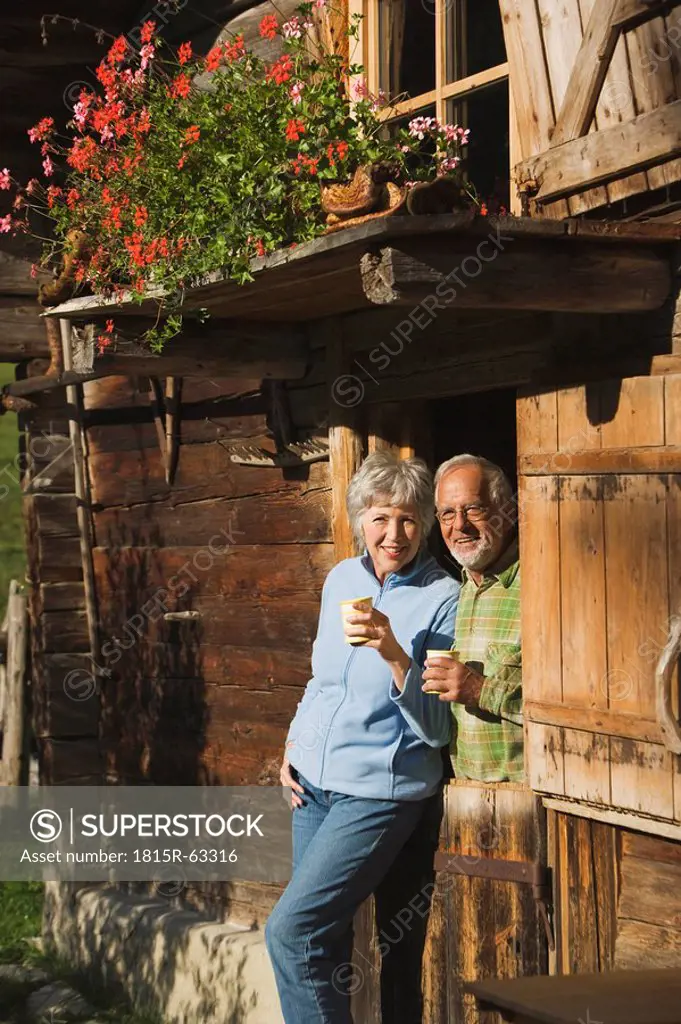 Austria, Karwendel, Senior couple leaning on log cabin, holding mugs