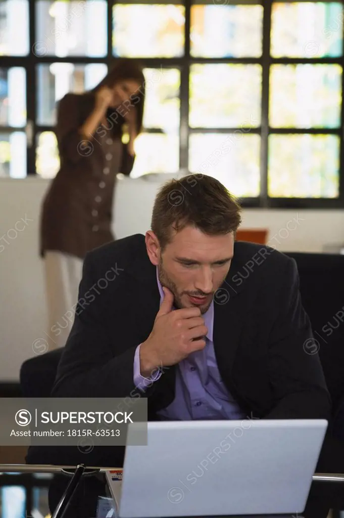 Man in office using laptop, woman in background phoning
