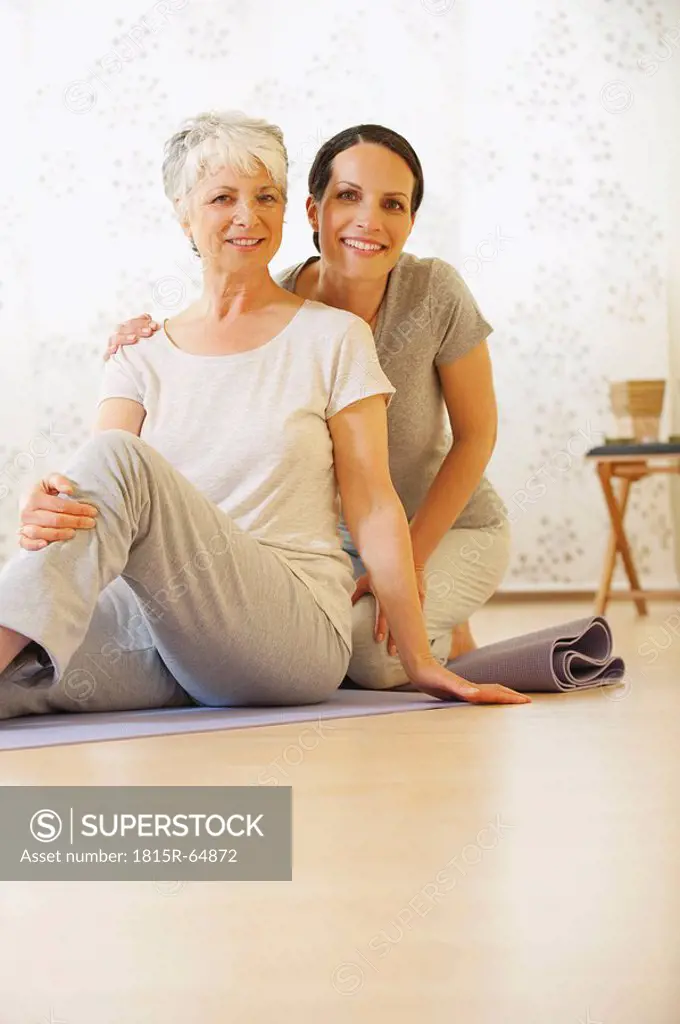 Two women sitting on gym mat, smiling, portrait