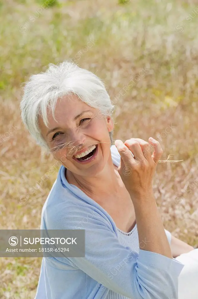 Spain, Mallorca, Senior woman sitting in meadow, holding blade of grass, laughing, elevated view