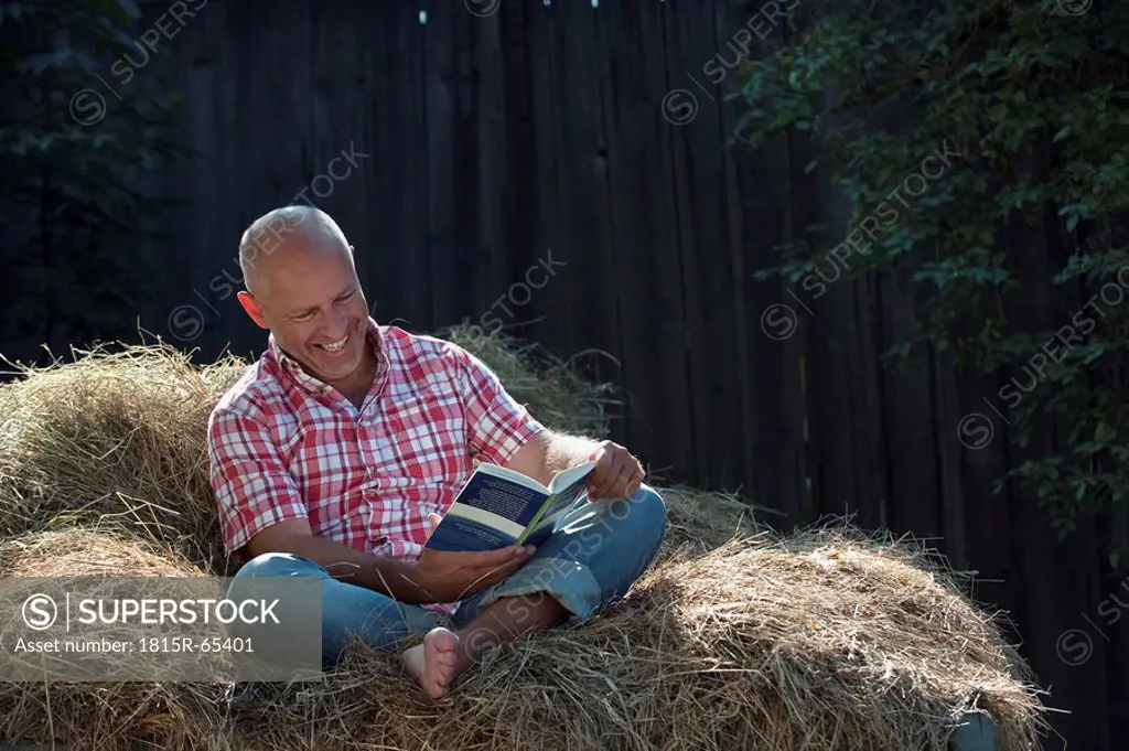 Germany, Bavaria, Senior man sitting on haystack, reading a book