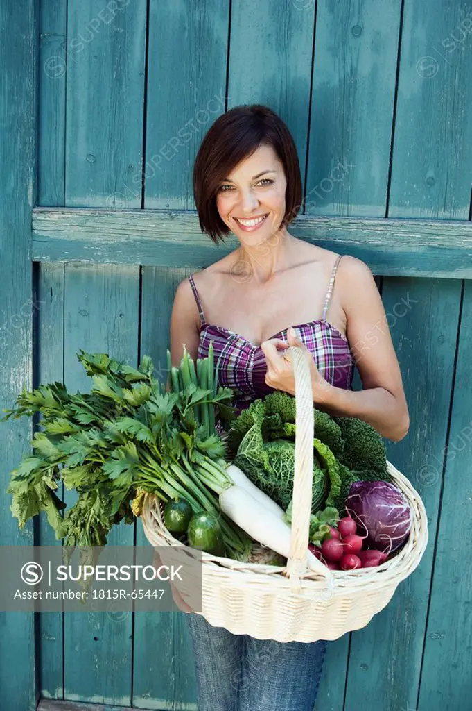 Germany, Bavaria, Woman in front of barn door holding basket with fresh vegetables, smiling, portrait