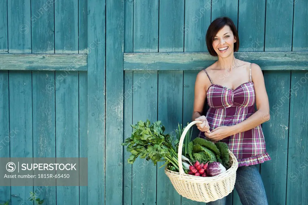 Germany, Bavaria, Woman standing in front of barn door, holding basket with fresh vegetables, smiling, portrait