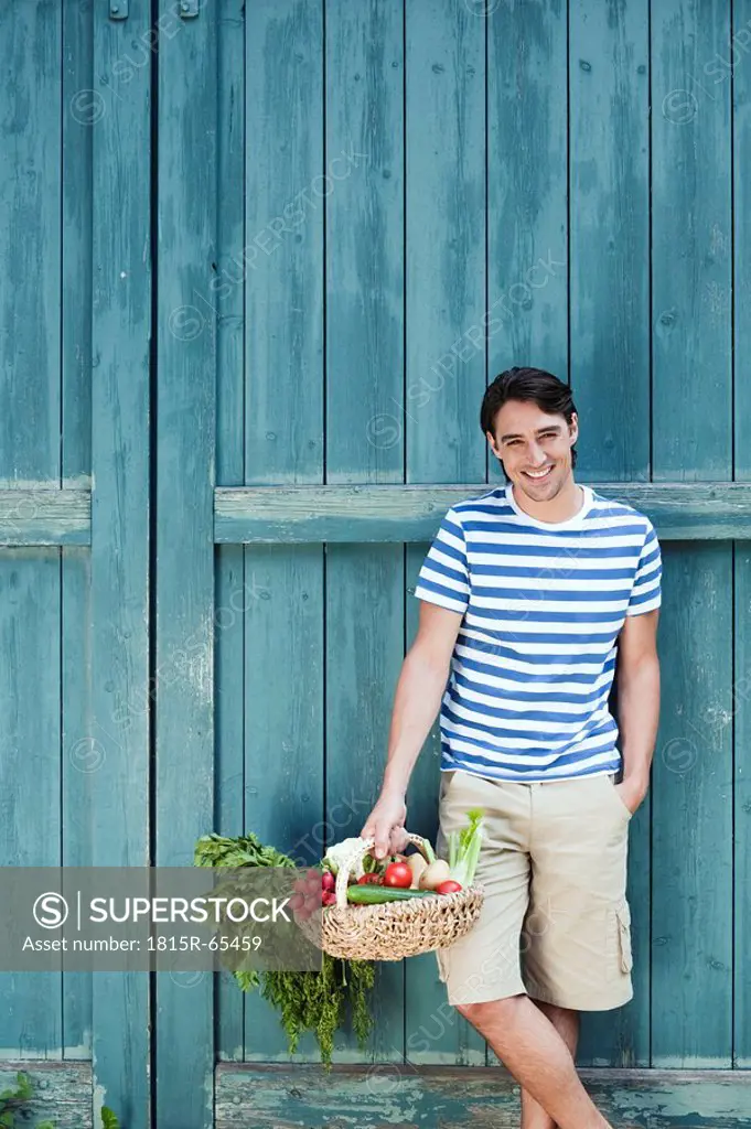 Germany, Bavaria, Man in front of barn door holding basket with fresh vegetables, smiling, portrait
