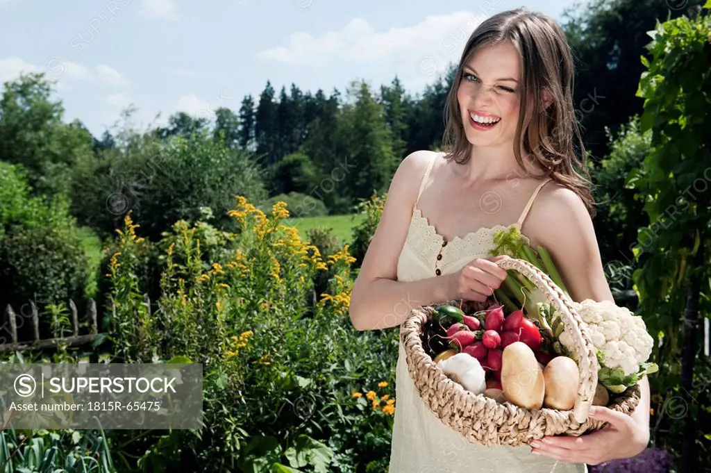 Germany, Bavaria, Young woman holding basket of vegetables in garden, smiling, portrait