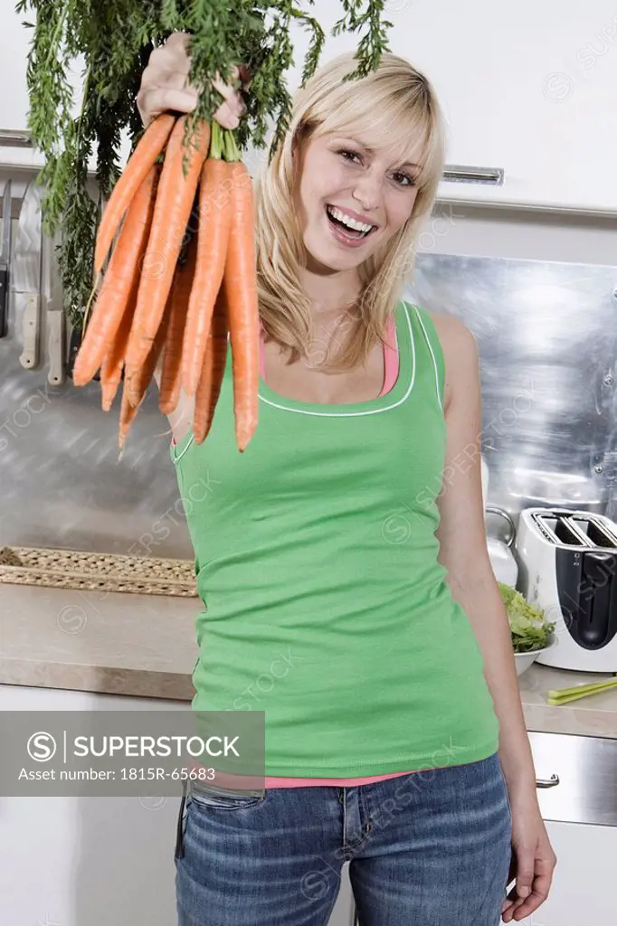 Germany, Berlin, Young woman in kitchen holding bunch of carrots, portrait