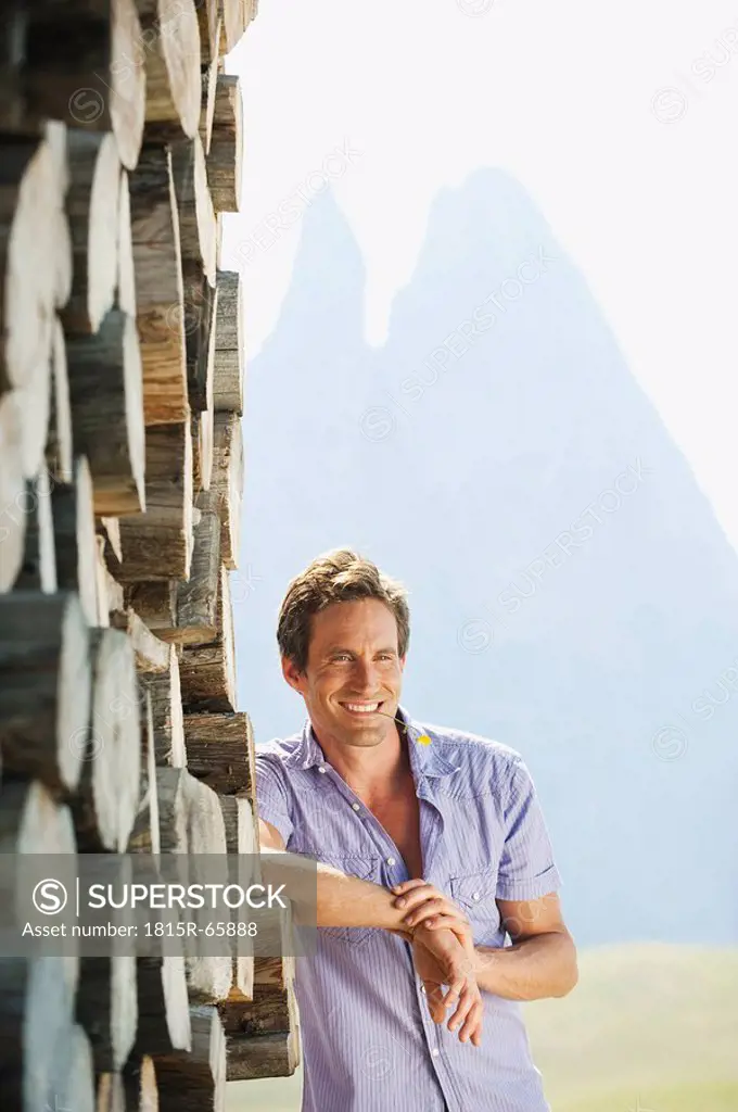 Italy, South Tyrol, Seiseralm, Man leaning against pile of wood, flower in mouth, smiling, portrait
