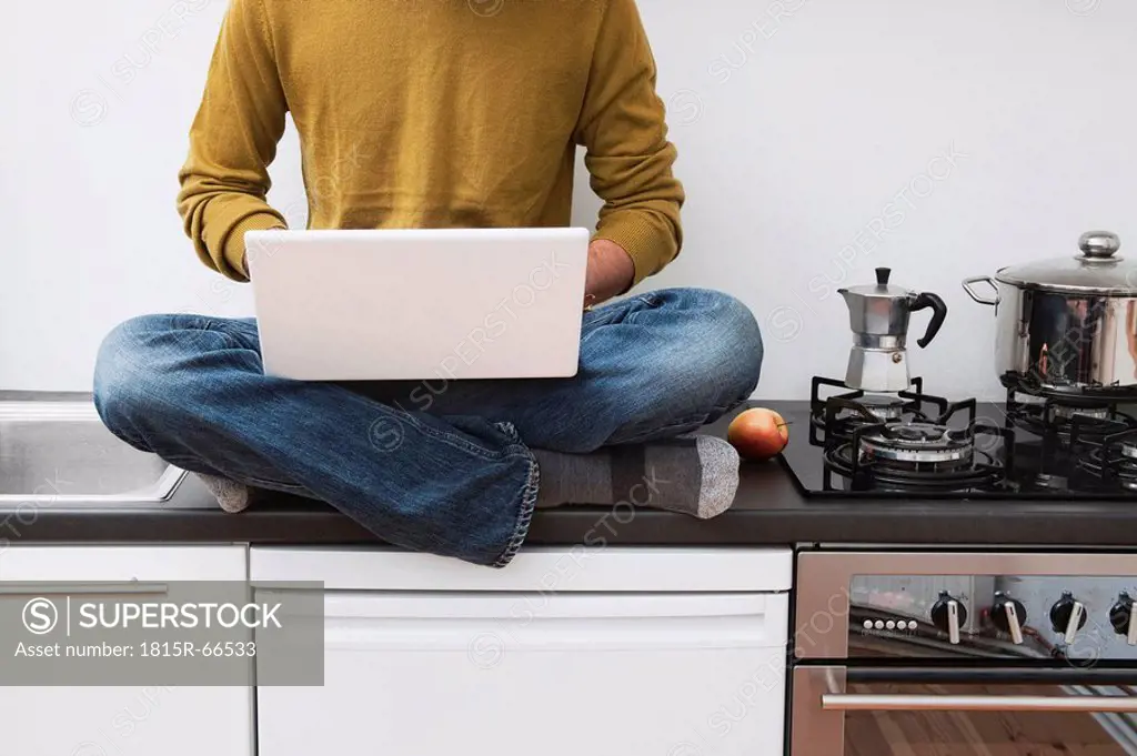 Man sitting on kitchen worktop, using laptop