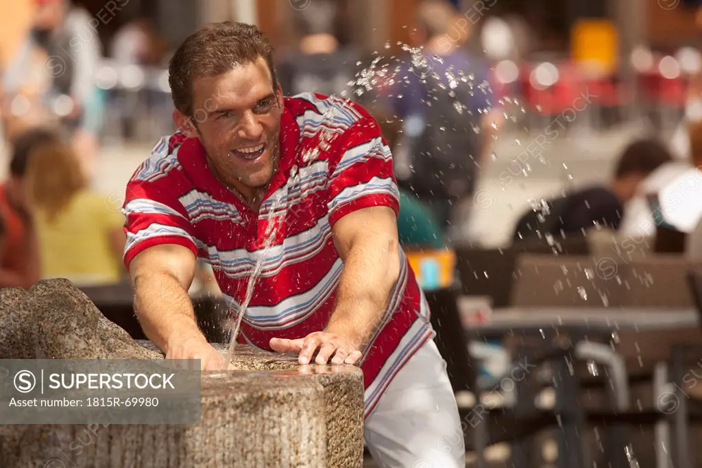 Germany, Bavaria, Regensburg, Man playing with fountain water, background people