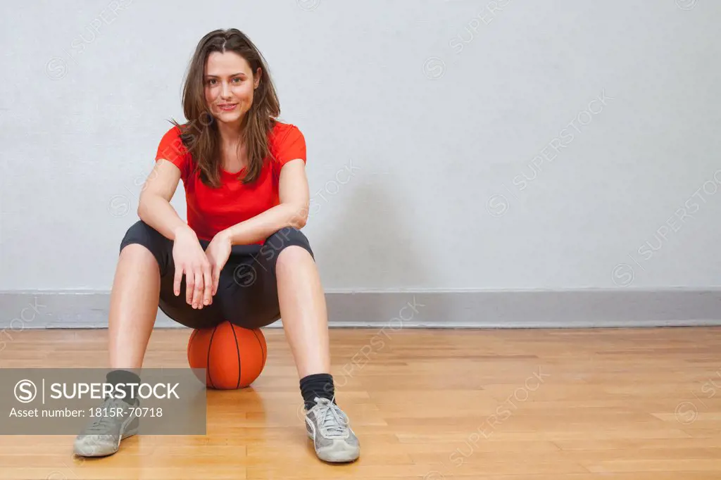 Germany, Berlin, Young woman sitting on basketball in school gym, smiling