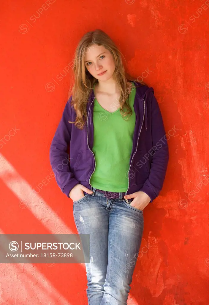 Europe, Teenage girl leaning and standing against red wall, smiling, portrait