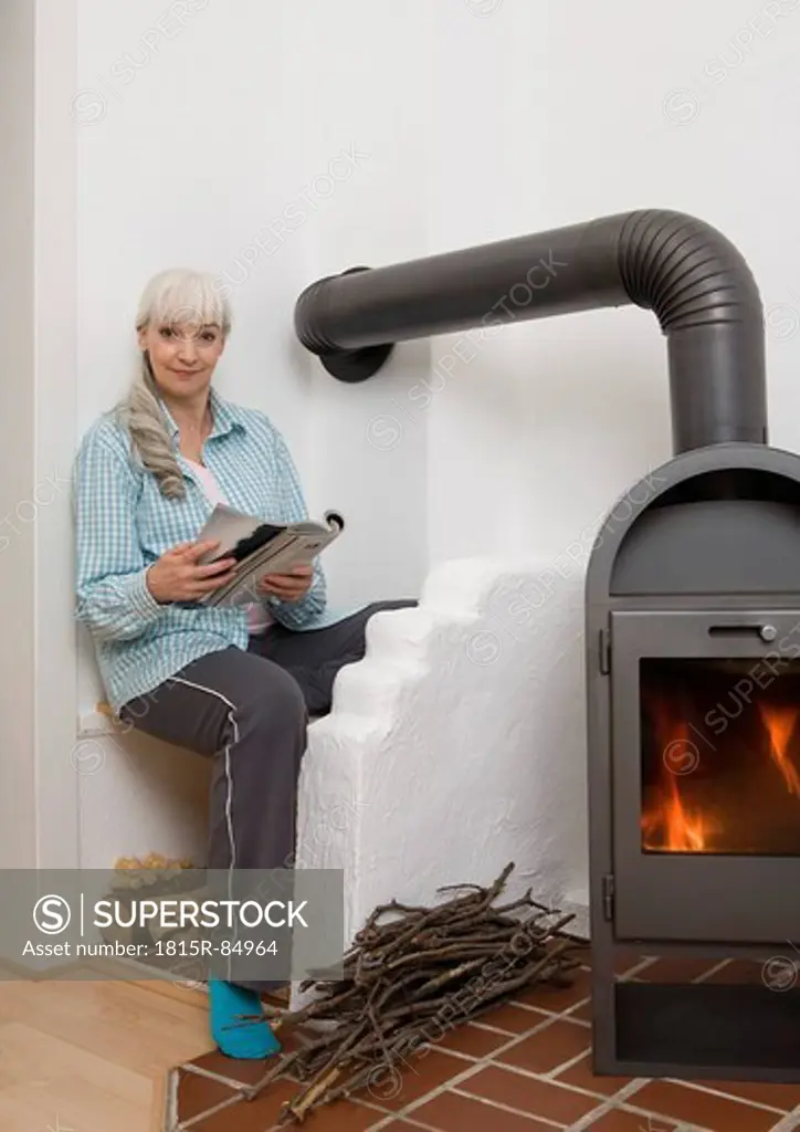 Germany, Duesseldorf, Woman sitting with magazine near fireplace, smiling, portrait