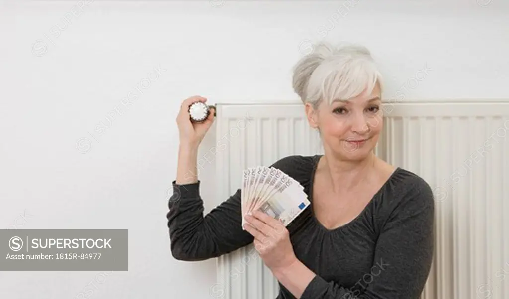 Germany, Duesseldorf, Woman holding banknotes and adjusting heater at home, smiling, portrait