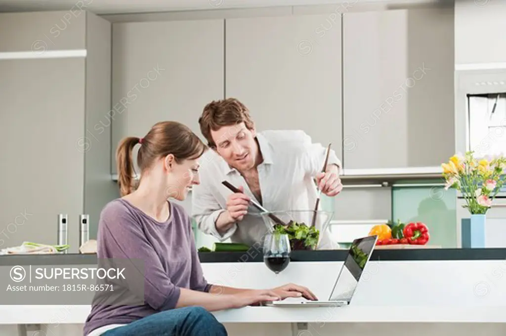 Germany, Hamburg, Man preparing salad with woman using laptop in kitchen