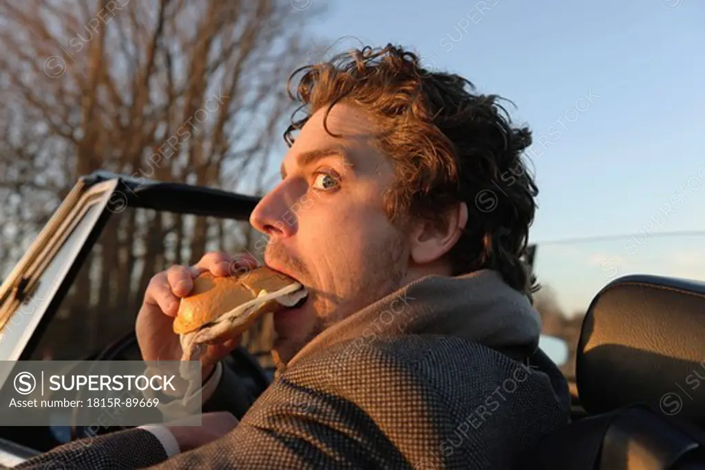Germany, Hamburg, Man eating fish sandwich in classic cabriolet car