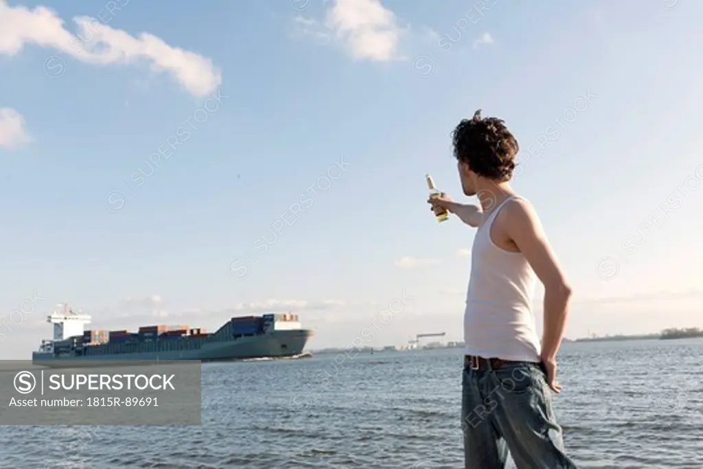 Germany, Hamburg, Man with beer bottle near Elbe riverside and container ship in background