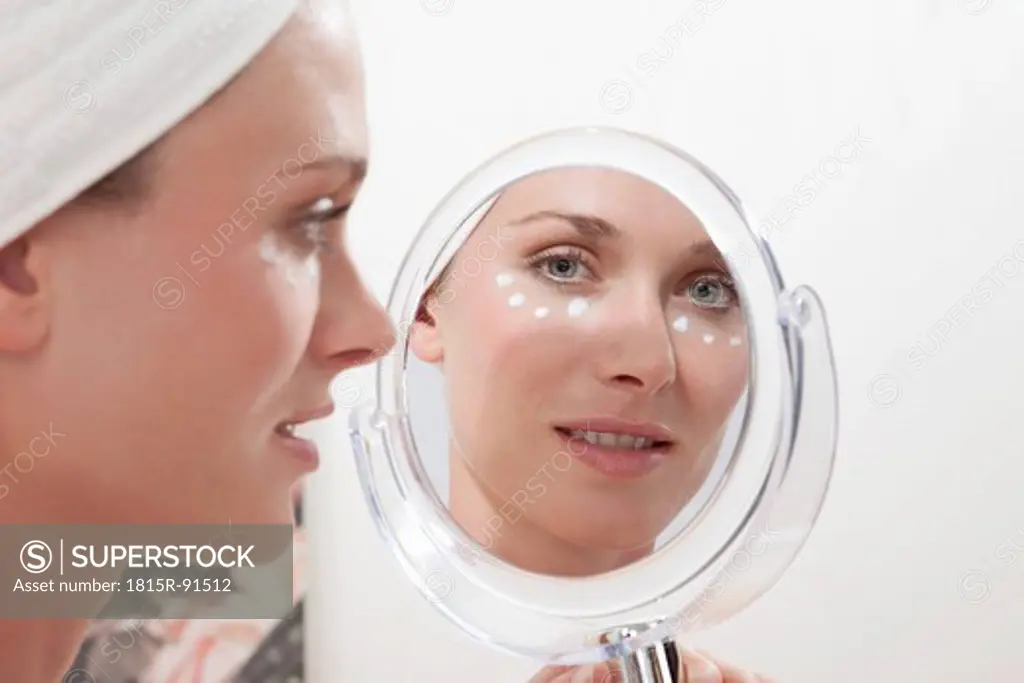 Young woman wrapped in towel appyling face cream and looking in mirror