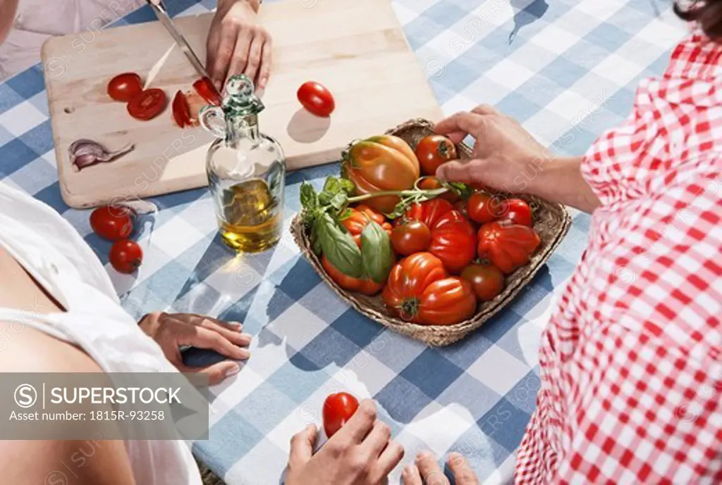 Italy, Tuscany, Magliano, Young woman cutting tomatoes on table with friends in foreground