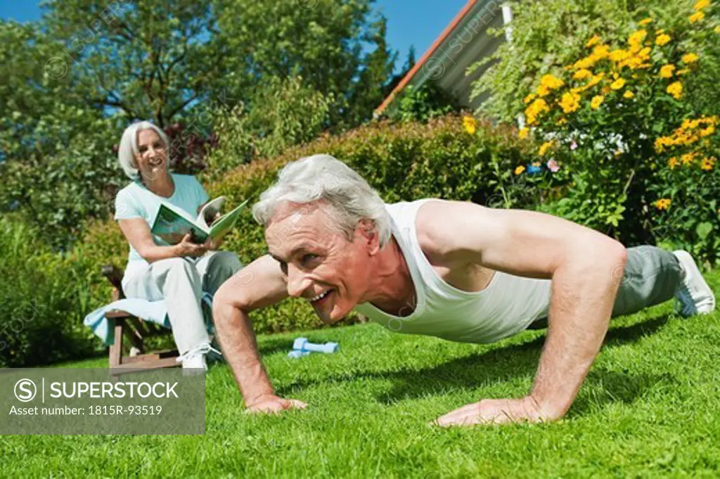 Germany, Bavaria, Man exercising and woman reading magazine in garden, smiling