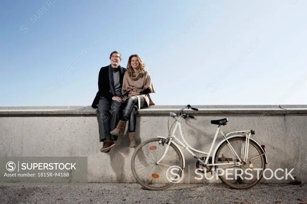 Germany, Bavaria, Munich, Young couple sitting on wall with bike