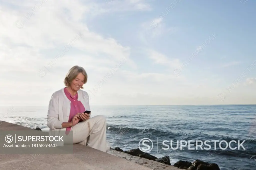Spain, Mallorca, Senior woman sitting and using mobile phone at sea shore, smiling